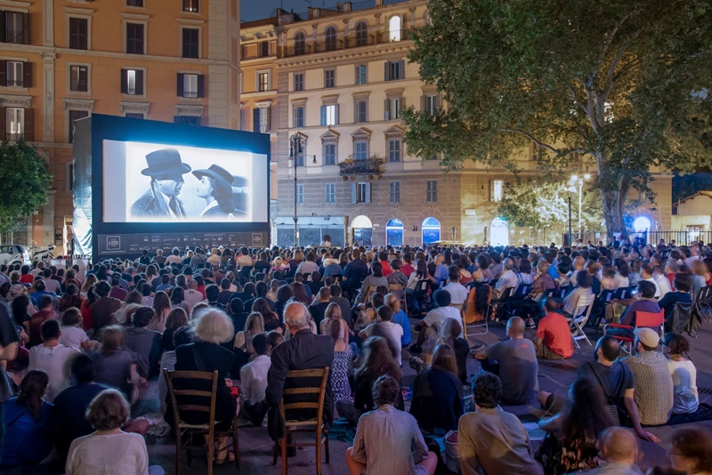 Outdoor cinema in a Rome piazza on a warm August evening, with people watching a movie under the stars